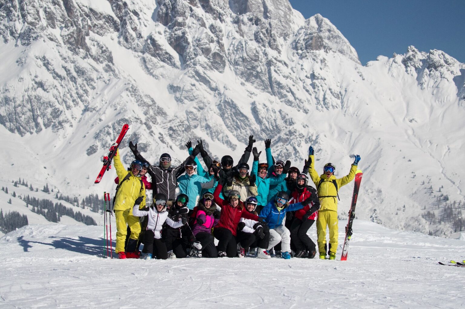 Eine jubelnde Gruppe aus 12 Skifahrerinnen und Skifahrern vor einen Bergkulisse in Skiausrüstung am Ende der Ausbildung.